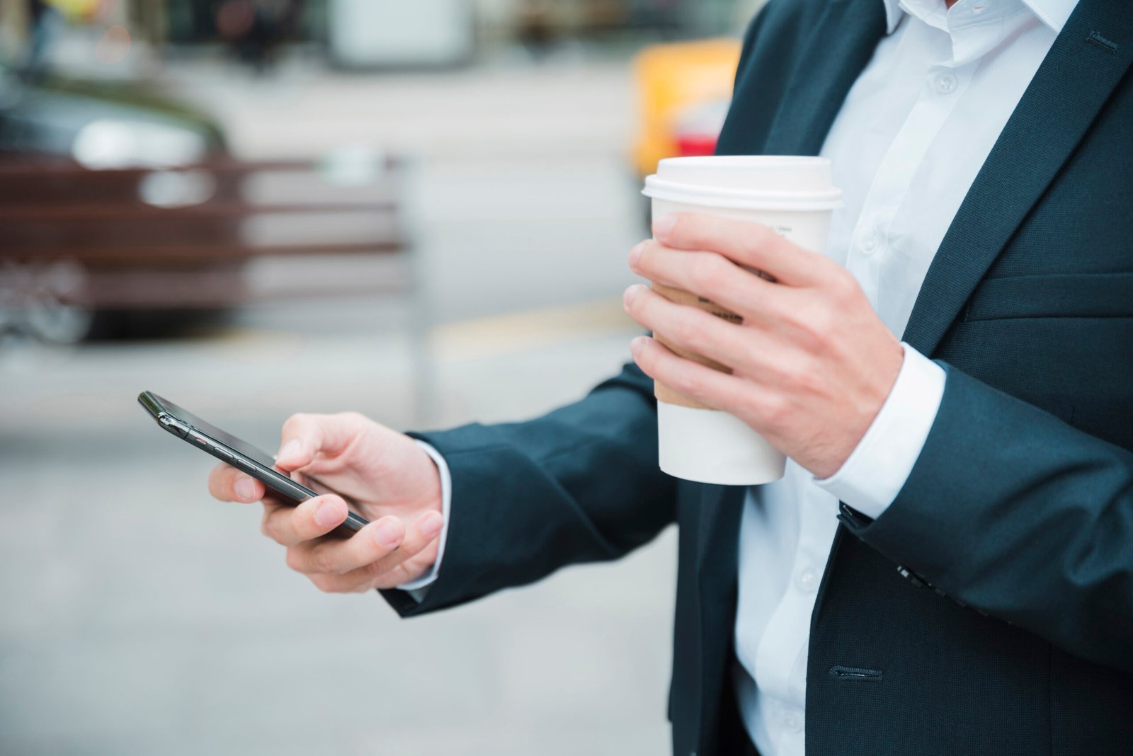 close-up-businessman-s-hand-holding-takeaway-coffee-cup-using-mobile-phone